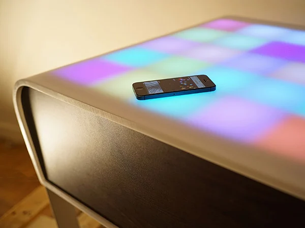 TA+BLE, table basse à cases lumineuses multicolores, avec un iPhone posé sur le plateau.