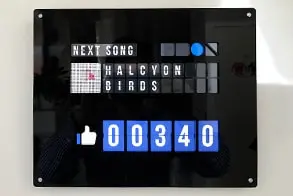 Un boîtier noir affiche un compteur de likes lumineux et le titre du prochain morceau.